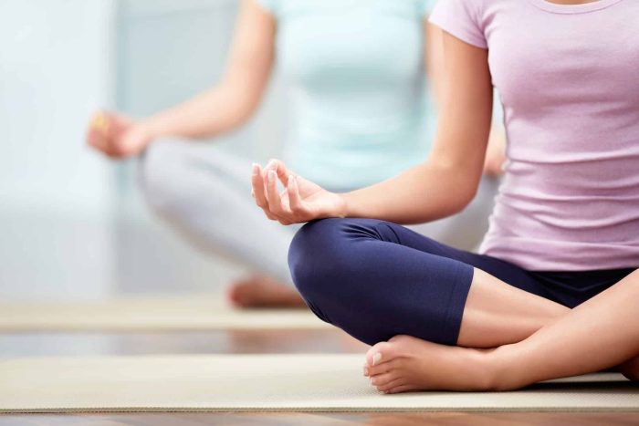 Two individuals practice yoga in a calm setting, sitting cross-legged on mats. The closer person wears a pink shirt and navy leggings, with hands resting on knees in a meditative gesture. The background person is less distinct, wearing light-colored clothing.