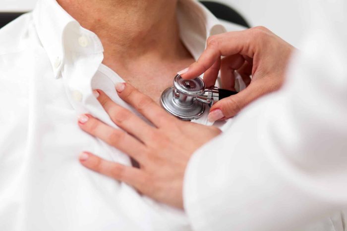 A healthcare professional uses a stethoscope to listen to a patient's heartbeat. The patient is wearing a white shirt, and the stethoscope is placed on their chest. The professional's hands are gently holding the stethoscope.
