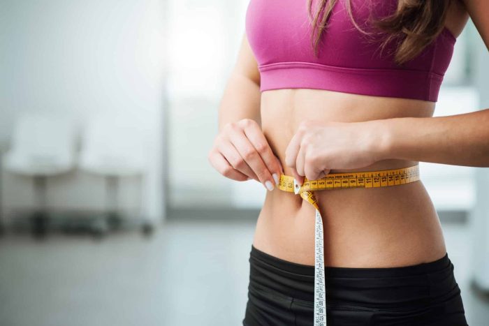 A woman measuring her waist with a yellow tape measure. She is wearing a pink sports bra and black leggings. The background is out of focus, showing some white, minimalistic furniture.