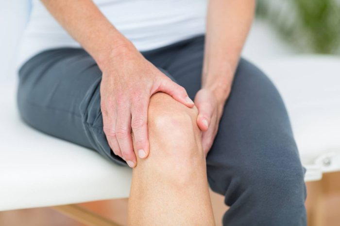 Close-up of a person wearing grey pants and a white top, sitting and holding their left knee with both hands. The individual appears to be comforting or examining the knee, suggesting possible discomfort or pain. The background is blurred and indistinct.