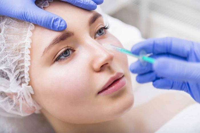 A person with a white hair cap receives a cosmetic injection in the nose area. A healthcare professional wearing blue gloves administers the injection using a syringe. The person appears calm with relaxed facial expression.