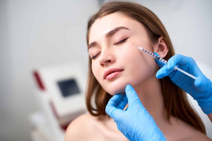 A person with closed eyes is receiving a cosmetic injection on their cheek from a gloved hand. The setting appears to be a clinical environment with medical equipment in the background. The individual looks relaxed and composed.