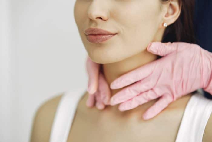 A person wearing pink gloves gently touches a woman's neck, likely preparing for a non-invasive neck lift. The woman, dressed in a white top, has only her lower face and neck visible in the image.