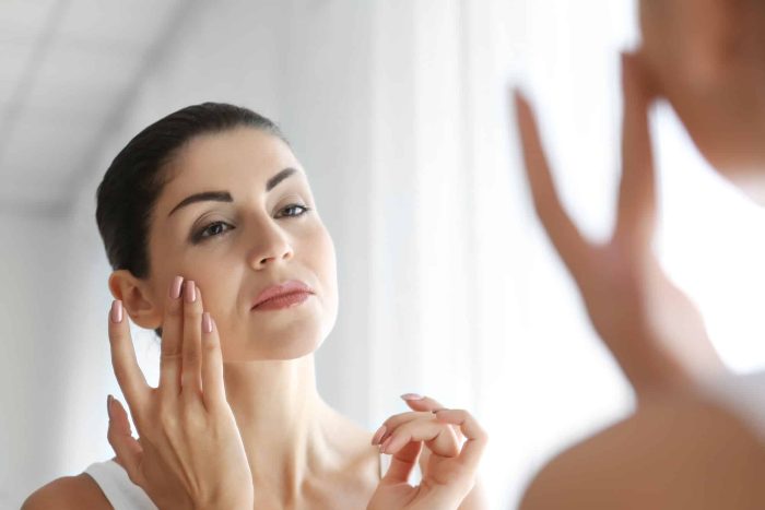 A woman with dark hair is standing in front of a mirror, gently touching her face with her fingertips. She appears to be examining her skin. The image has a bright, soft, and natural lighting, enhancing the serene and focused atmosphere.