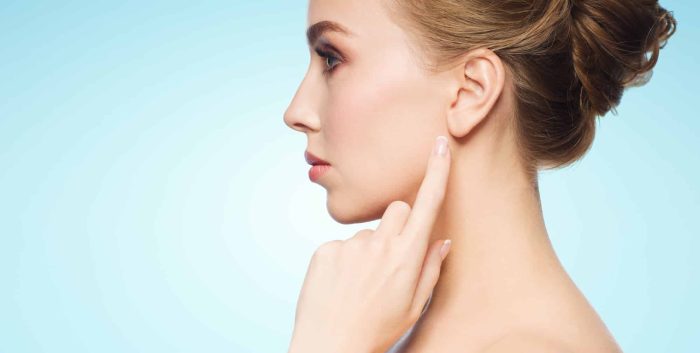 A woman with tied-up hair points to her ear with her index finger. She is seen in profile against a light blue background. The image focuses on her clear complexion and features a serene expression.