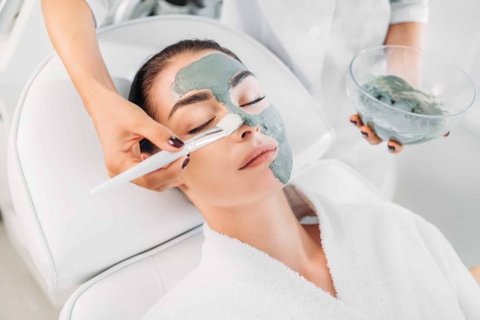 A woman is relaxing in a spa while a beautician applies a green facial mask to her face with a brush. The woman is wearing a white robe and reclined on a comfortable chair. The beautician holds a bowl of the mask in one hand.