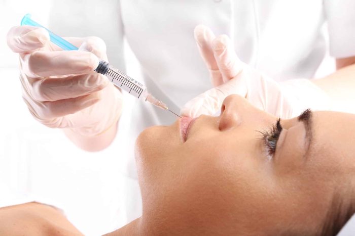 A healthcare professional wearing gloves holds a syringe near a patient’s lips, preparing to administer an injection. The patient lies down with eyes closed, appearing relaxed. This setting suggests a cosmetic or medical procedure.