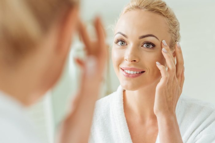 A woman with blonde hair is looking into a mirror and gently touching her face with a smile. She is wearing a white bathrobe and appears to be in a well-lit, clean bathroom. The focus is on her reflection, showing her engaging in a skincare routine informed by her recent DNA skin test results.