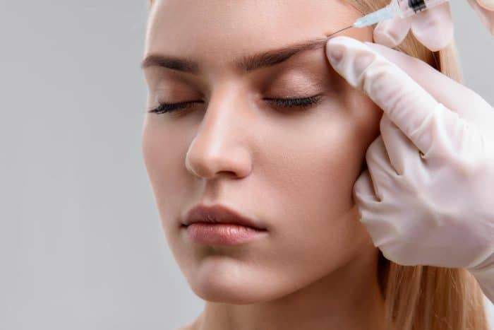 A close-up of a person with closed eyes receiving a botox brow lift in their forehead. They have light skin and blonde hair. The one administering the injection is wearing white gloves. The background is neutral and out of focus.