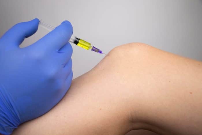 A close-up of a person's knee receiving an Orthovisc injection. A gloved hand is holding a syringe and administering the shot. The background is out of focus, highlighting the Orthovisc injection procedure on the knee.