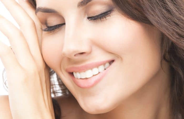 A close-up of a smiling person with long, brown hair and closed eyes. They are gently touching their face with one hand, displaying a serene and content expression. The background is a light, neutral color, enhancing the softness and warmth of the image, perfectly capturing the beauty of their gummy smile.
