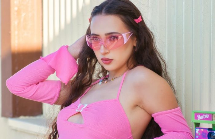 A person with long hair stands outdoors, wearing pink-tinted sunglasses and a chic pink outfit featuring a top with a keyhole detail. They have a confident expression, posed with one hand behind their head. Surrounded by pink accessories and a Barbie box, they exude an effortless, barbie-like charm.