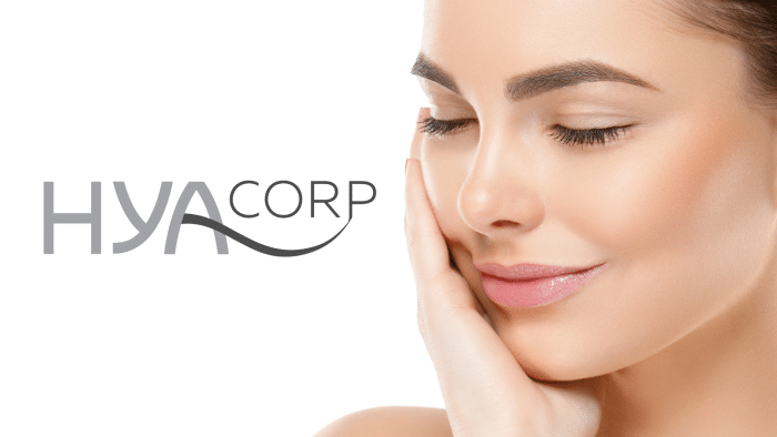 A close-up of a woman with clear skin touching her cheek with a gentle smile, eyes closed, showcasing a stunning hyacorp before and after transformation. The logo "HYA CORP" is displayed on the left against a white background.