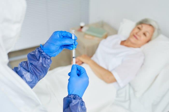 A healthcare worker in protective gear prepares a syringe for Orthovisc injections for knee pain, while an elderly woman lies in bed in the background.