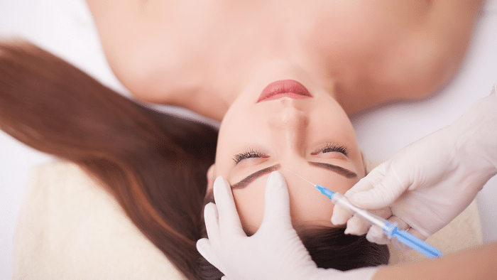 A person lies down with eyes closed while receiving a cosmetic procedure on their forehead. A practitioner with gloved hands is administering an injection with a syringe. The person has long, brown hair spread out on the pillow, pondering how long Botox can last after this session.