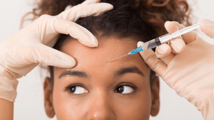 A close-up of a person receiving a cosmetic injection in the forehead. The person, curious about what age to start Botox, has curly hair and is looking slightly upwards. The hands performing the injection are wearing white gloves and holding a syringe with a blue needle aimed at the person's forehead.