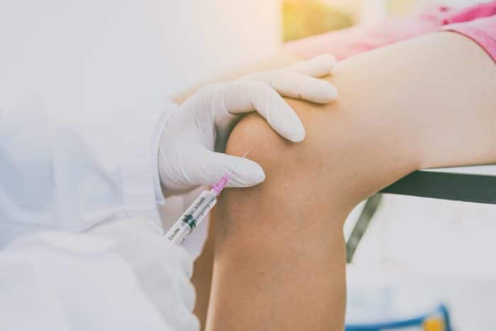 A medical professional wearing gloves administers an injection into a patient's knee, perhaps addressing concerns about how long Orthovisc lasts. The patient is seated, with one gloved hand stabilizing the knee while the other hand holds a syringe. The background suggests a clinical setting.
