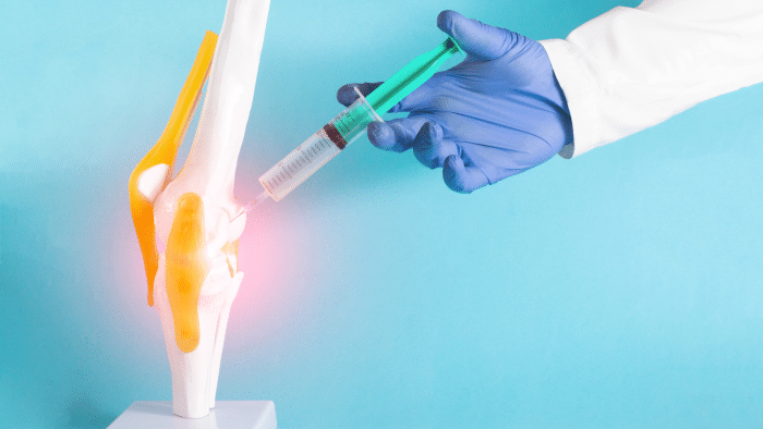 A model of a human knee joint is shown against a light blue background. A hand in a blue glove holds a syringe, administering Orthovisc treatment into the knee joint. The knee has yellow ligaments, and a red glow indicates pain or inflammation around the injection site.