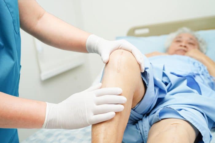 A healthcare professional wearing gloves examines an older patient's knee, which has a visible scar, likely discussing orthovisc injection aftercare. The patient is lying on a hospital bed, partially covered with a blue sheet, in a medical setting.