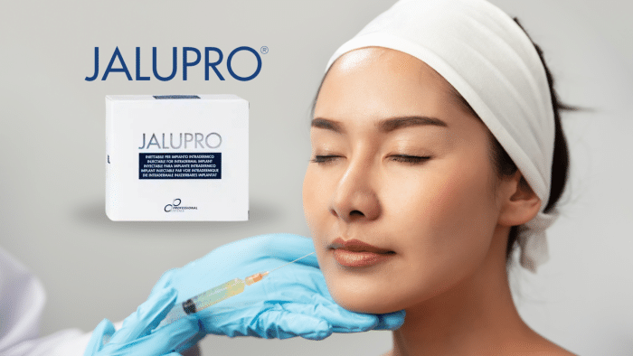 A doctor wearing blue gloves is injecting a woman's upper cheek with a syringe. The woman has her eyes closed and looks calm. The image includes a box of Jalupro, highlighting the Jalupro benefits for skin treatments, with the Jalupro logo in the background.