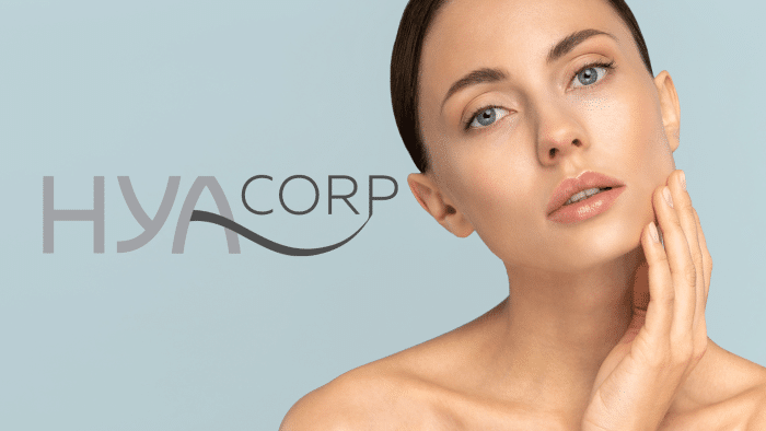 A woman with clear skin and minimal makeup touches her face gently with one hand. The image background is light blue, and the text "HYA Corp" is displayed beside her in a sleek font. She has a calm and serene expression, hinting at the serene effects of a Hyacorp injection.