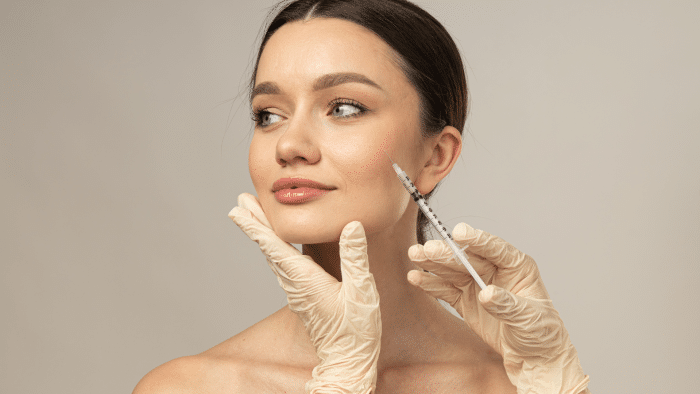 A woman with bare shoulders receives an injection in her cheek from a person wearing latex gloves. The woman has a calm expression and looks off-camera, exemplifying what Botox can do to your face. The background is neutral and plain.