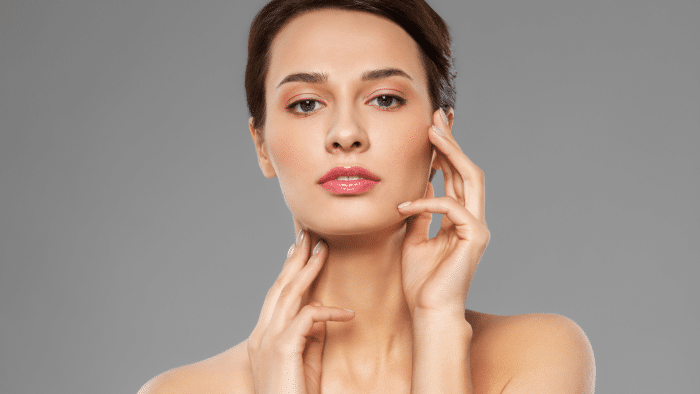 A woman with clear skin and neutral makeup poses against a gray background. She touches her face with her fingers, one hand near her chin and the other beside her cheek, exuding confidence and serenity—reflecting how Botox was discovered to enhance natural beauty.