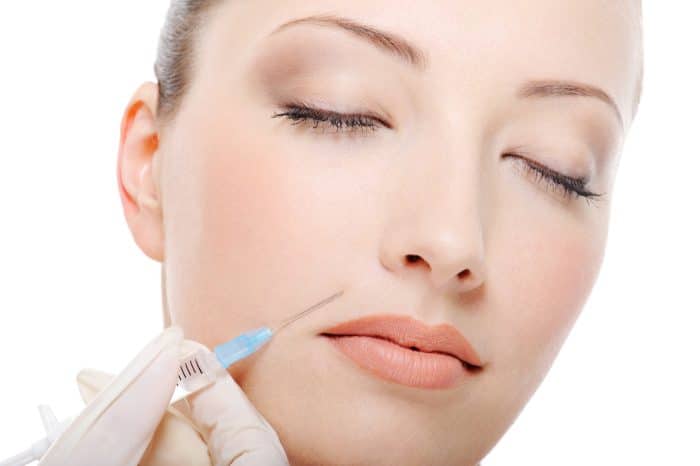 A close-up of a person's face with eyes closed while receiving a Dysport injection near the corner of their mouth. A gloved hand holds the syringe. The background is white.