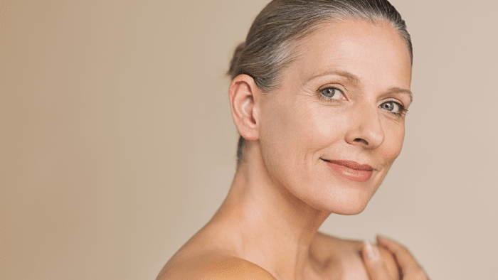 A senior woman with light skin and gray hair styled in a low bun smiles softly at the camera. She has a natural makeup look, reminiscent of someone who might choose Restylane Refyne vs Defyne, and is posed against a plain beige background, exuding a sense of calm and confidence.