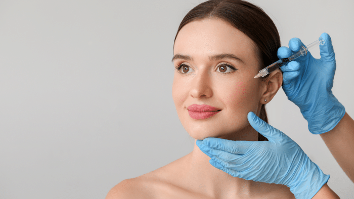 A woman with brown hair is receiving a Restylane injection on the side of her face. She is looking slightly upward and smiling gently. The procedure is being administered by gloved hands holding a syringe. The background is neutral and light gray.