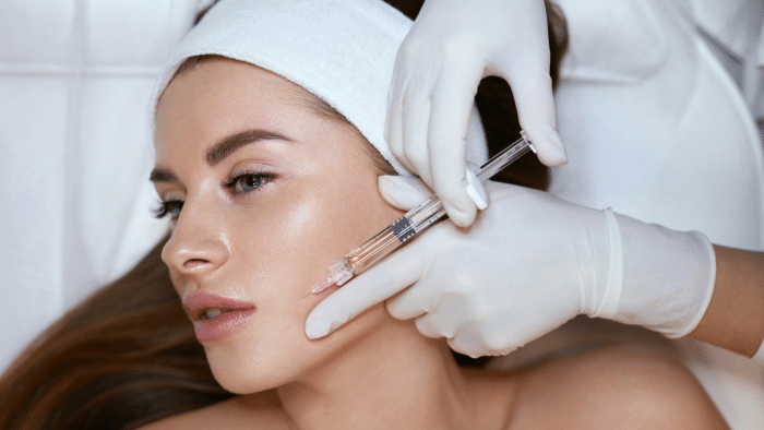 A woman is receiving a cosmetic injection in her cheek. She is lying down with her head resting on a white surface, and a person wearing white gloves is administering the injection, referencing the xeomin dosing chart. The woman has long brown hair and is wearing a white headband.