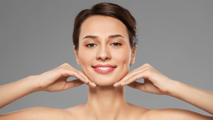 A person is posing against a gray background, smiling gently. They have dark hair pulled back and are holding their hands under their chin, with fingers lightly touching their jawline. The person is demonstrating a relaxed, confident expression, highlighting the impressive Xeomin results.