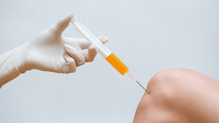 A gloved hand administers an orange liquid injection into the side of a person's bare knee against a plain white background, in accordance with Hyalgan prescribing information.
