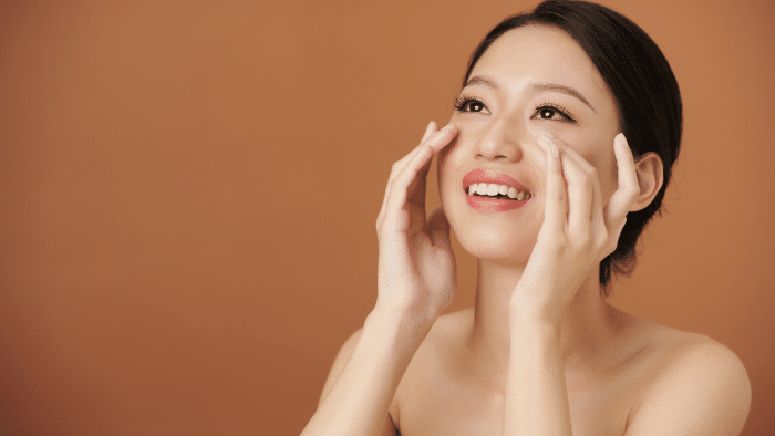 A woman with a joyful expression touches her cheeks with both hands. She has a bright smile, flawless skin, and dark hair pulled back. The background is a solid warm orange color, highlighting her radiant complexion.