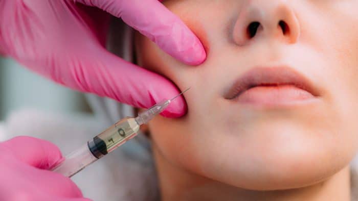 A close-up of a person's face receiving an injection in the cheek. The person performing the procedure is wearing pink gloves. The needle is close to the skin, and the syringe contains a clear liquid. The person's lips are slightly parted.