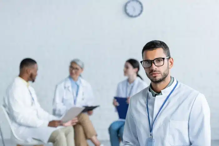 A group of medical professionals in white lab coats sits and discusses in the background, with a male doctor wearing glasses and a lab coat standing in the foreground, looking at the camera. A clock is on the white brick wall behind them.
