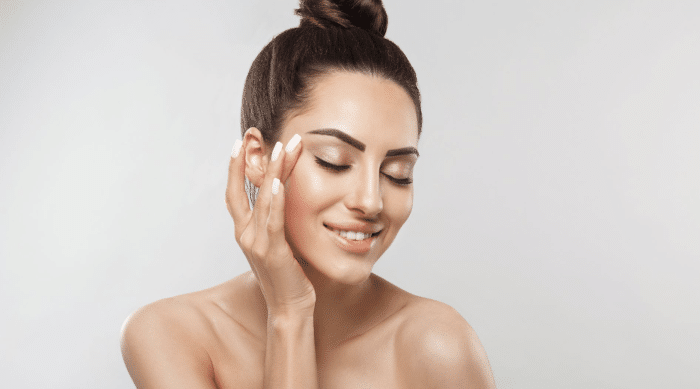 A woman with dark hair in a high bun is posing with her eyes closed and a slight smile. She is touching her face gently with one hand, showcasing her manicured nails. She has clear skin and is against a plain, light-colored background.