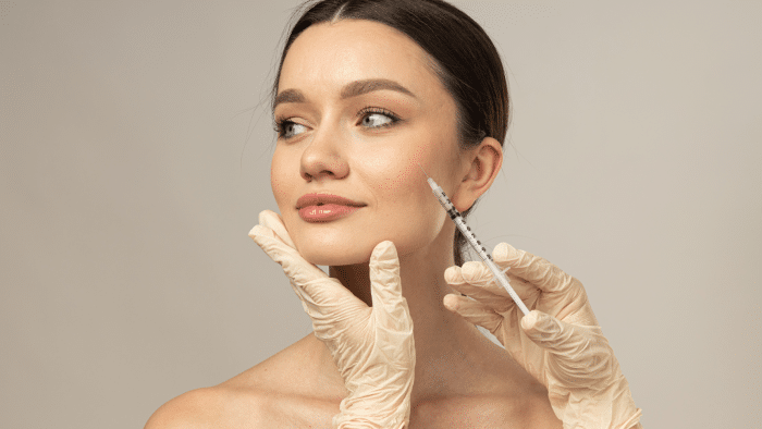 A woman with a serene expression receives a facial injection from a medical professional wearing latex gloves. The medical professional uses one hand to support her chin while holding the syringe with the other. The background is plain and light-colored.