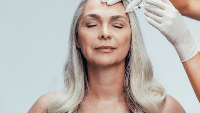 A woman with long, gray hair closes her eyes as she receives a cosmetic injection on her forehead from a gloved hand. The background is plain and light-colored, highlighting the procedure.