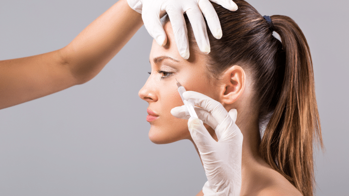 A woman with her hair in a ponytail is receiving a cosmetic injection in her face. She is looking calm as she is gently pulled by hands in white gloves. The background is plain, and the focus is on the woman's profile and the procedure.