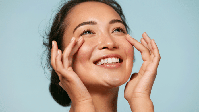 A woman with a radiant smile gently touches her face with both hands. She appears happy and content, looking upwards with a clear, bright complexion against a light blue background. Her hair is neatly pulled back.