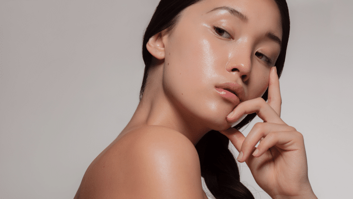 A person with clear, glowing skin is shown in a close-up portrait against a plain backdrop. They have long, dark hair pulled back and are resting their chin on their hand while gazing directly at the camera. Their expression is calm and introspective.
