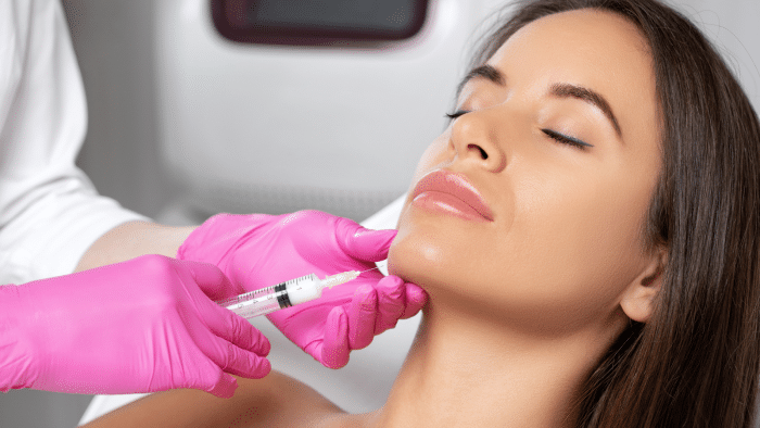 A person with long, dark hair and closed eyes is receiving a facial injection from another individual wearing pink gloves. The syringe is positioned near the person's chin. The background is blurred, focusing on the person and the procedure.