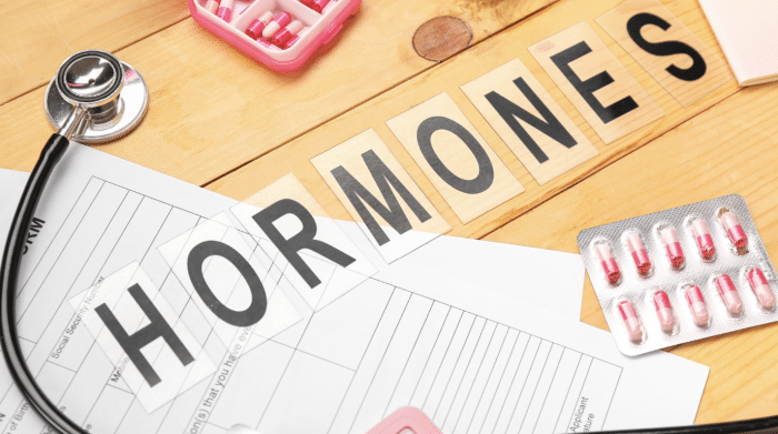 A wooden table with a stethoscope, medicine blister packs, a pill organizer, and documents. The word "HORMONES" is spelled out with letter tiles arranged on the table.