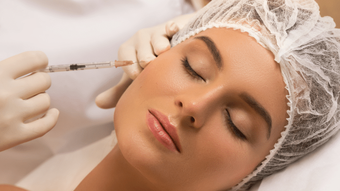 A person with closed eyes is receiving a facial injection from a gloved hand. The individual is wearing a white hair cap and appears relaxed. The background is softly blurred, focusing attention on the procedure.