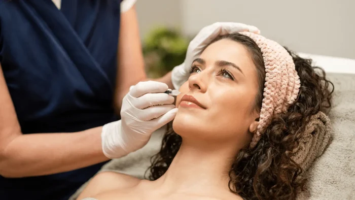 A woman with curly hair and a pink headband lays on a padded surface while receiving a cosmetic procedure. A professional wearing white gloves applies treatment to her lips. The setting appears clean and professional.