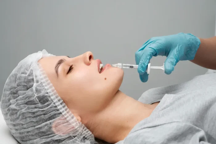 A person is lying down with their eyes closed, wearing a hair cap. They are receiving an injection near their chin from someone wearing blue gloves. The setting appears clinical or medical.