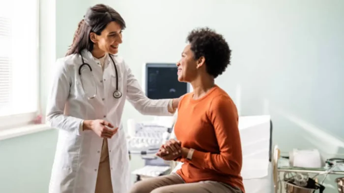 A doctor in a white coat smiles and converses with a seated patient in an orange sweater. The setting appears to be a medical office with equipment visible in the background. The mood is friendly and reassuring.