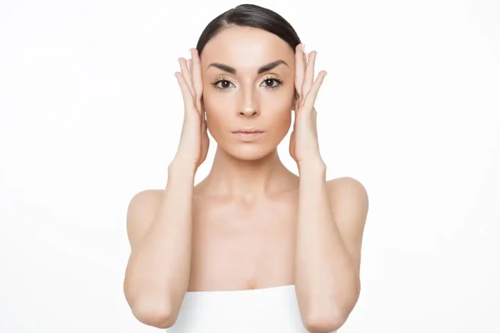 A person with dark hair and minimal makeup stands against a white background, holding their hands to their head. Their expression is neutral, and they are wearing a strapless top.