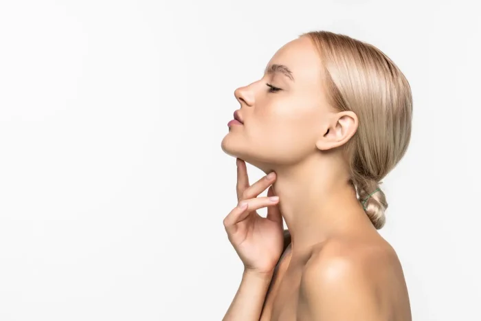 A young woman with blonde hair tied back, posing in profile against a white background, gently touching her chin and neck with her fingers.